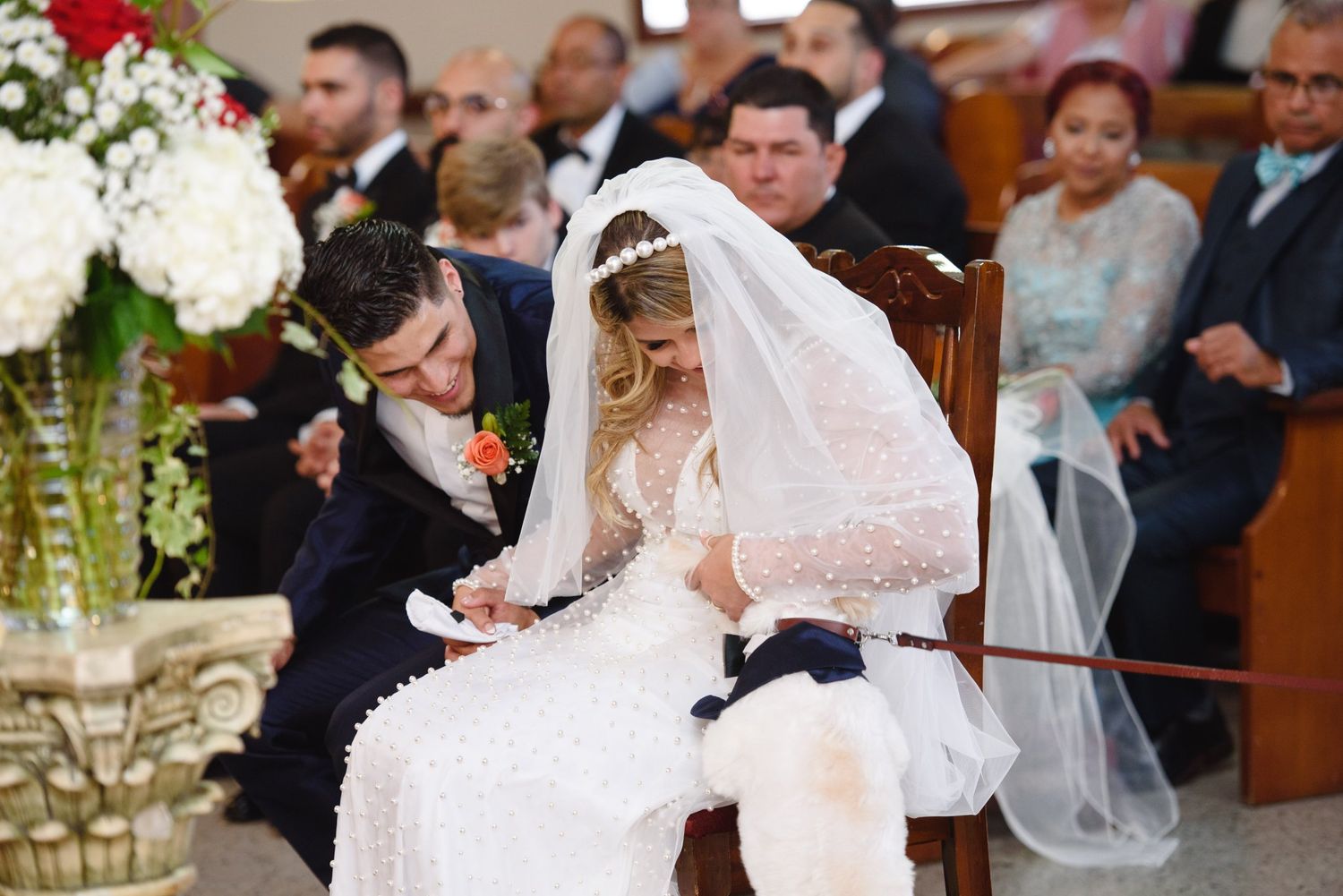 Bride and groom signing wedding documents at church ceremony, bride in lace gown and veil, guests seated in pews behind them.