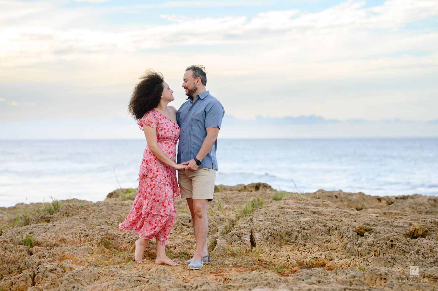 Couple holding hands on rocky coastal cliffs with ocean and cloudy sky backdrop.