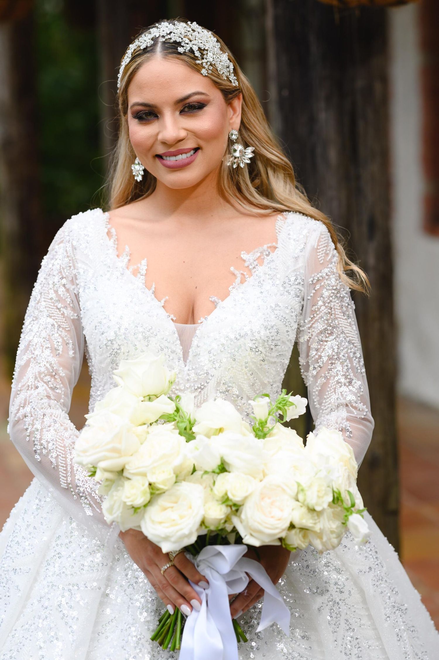 Smiling bride in beaded lace gown with sheer sleeves holds white rose bouquet, wearing crystal headpiece and earrings.