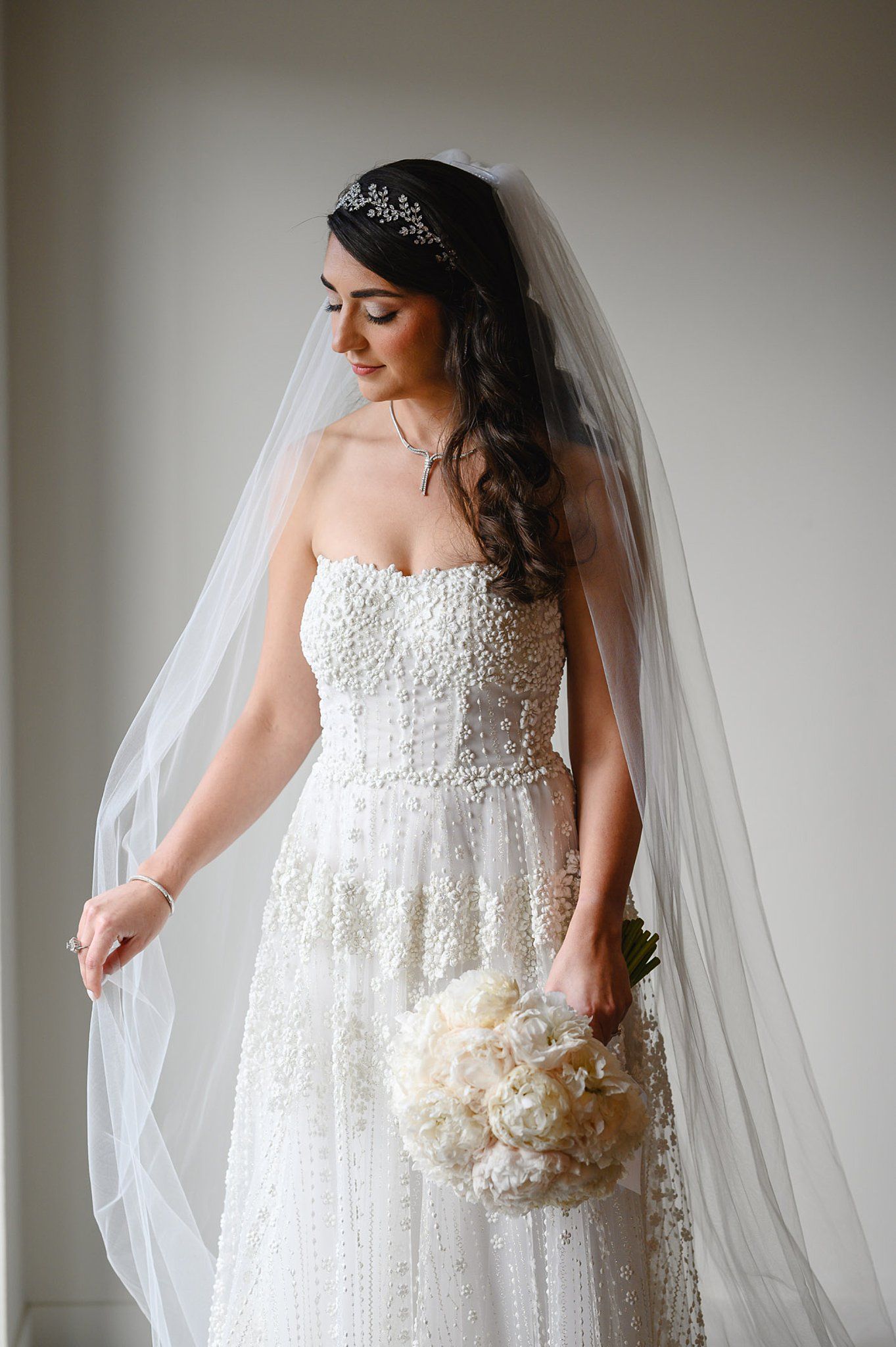 Bride in beaded strapless wedding gown with veil and crystal tiara holding white peony bouquet.