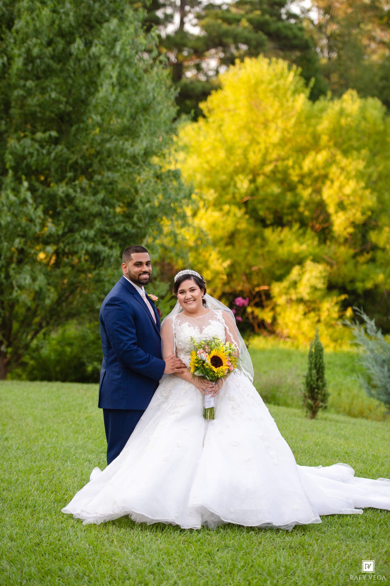Bride and groom pose outdoors in lush garden, she holds sunflower bouquet in white gown, he wears navy suit.