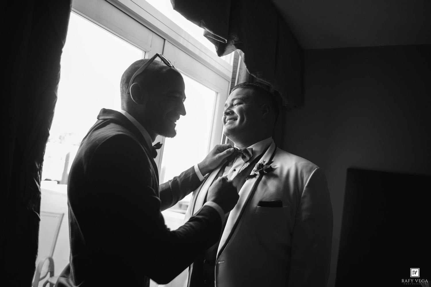 Black and white photo of a man helping a groom adjust his bow tie before a wedding ceremony.