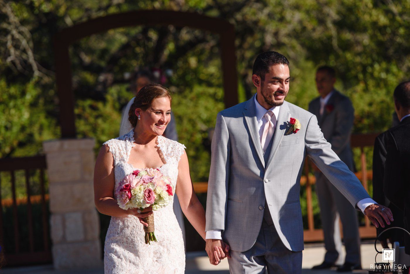 Smiling bride and groom walk hand-in-hand after outdoor wedding ceremony, bride holding pink bouquet.