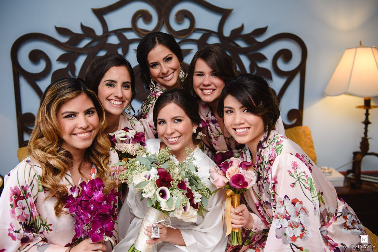 Bride in white robe holding bouquet surrounded by bridesmaids in floral robes before wedding ceremony.