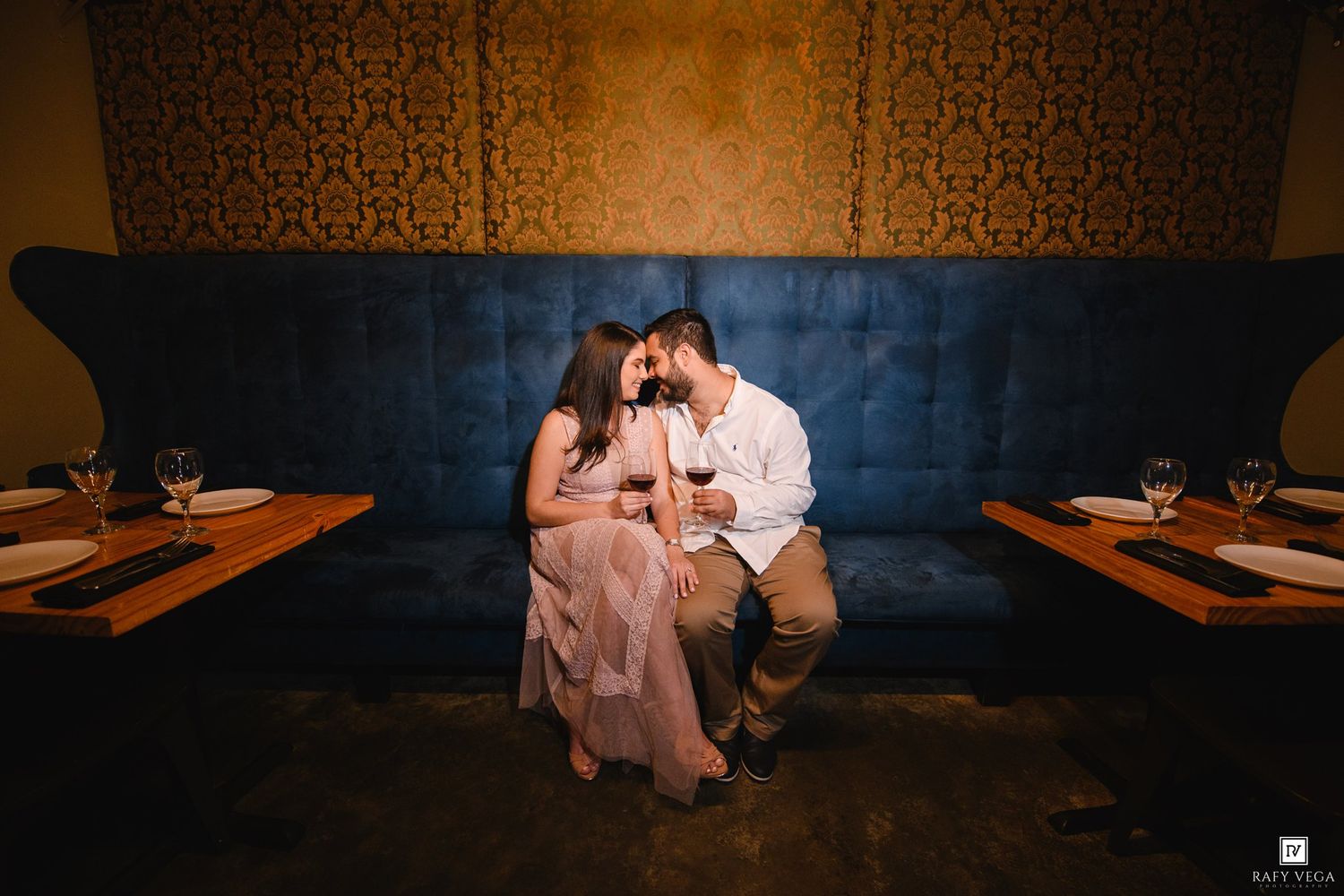 Romantic couple sharing wine on a blue velvet booth in an intimate dimly lit restaurant setting.