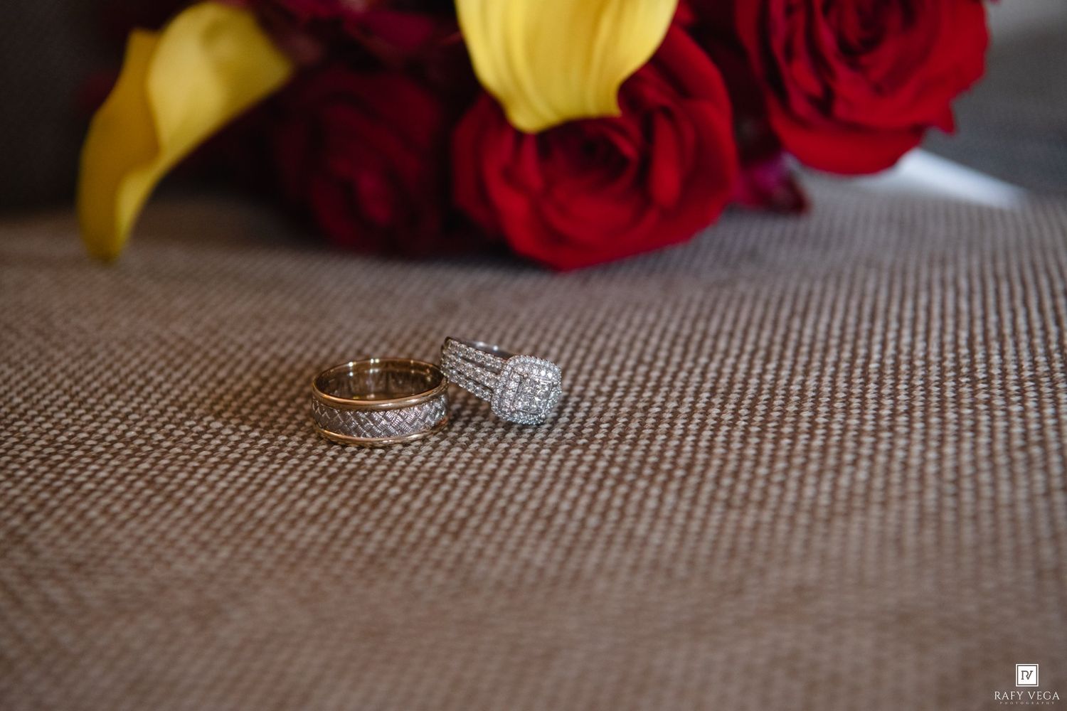 Gold wedding bands and diamond engagement ring displayed on burlap near red roses and yellow lilies bouquet.