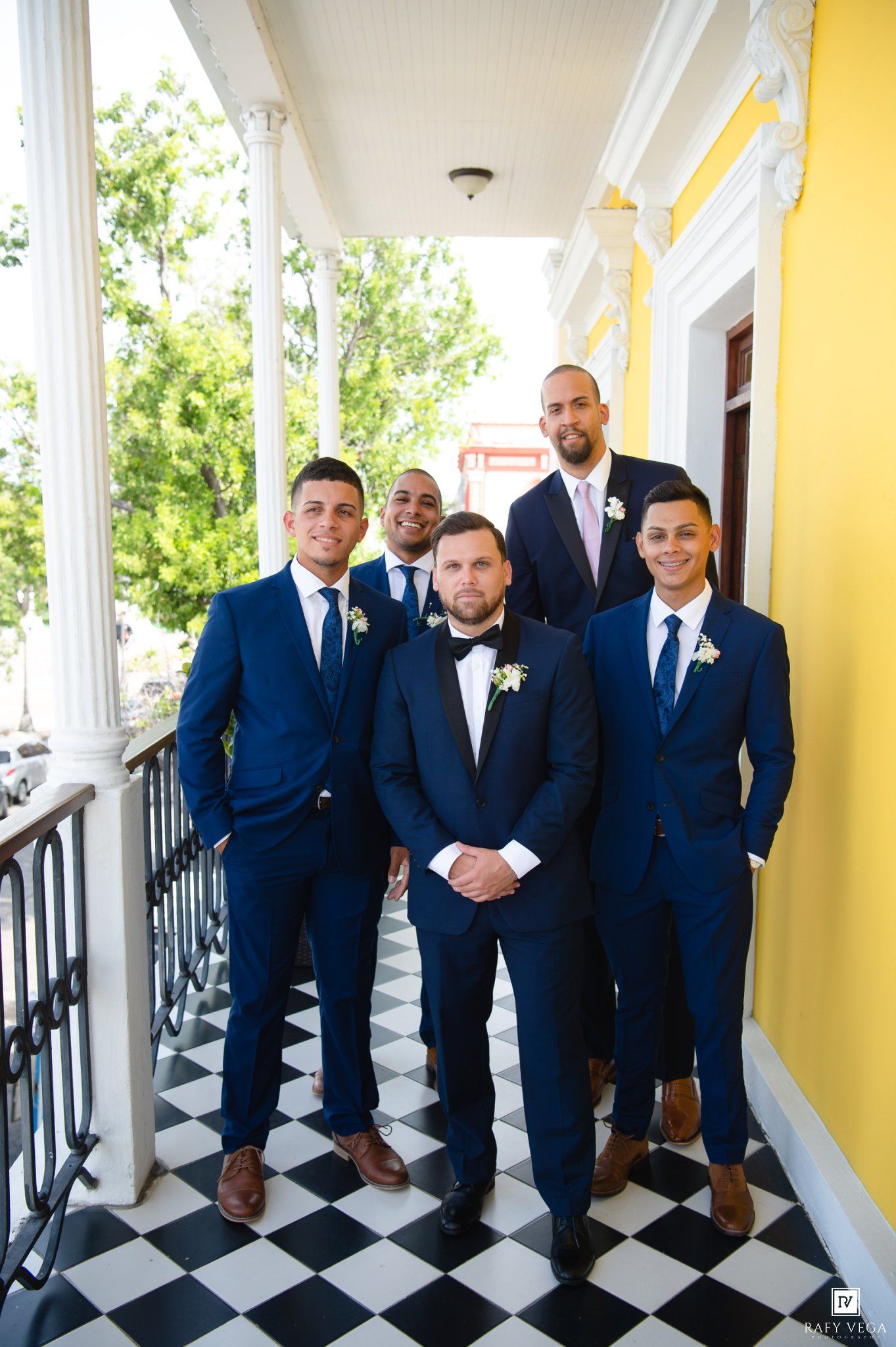 Groom and groomsmen in navy blue suits pose on a checkered floor balcony of a yellow building.