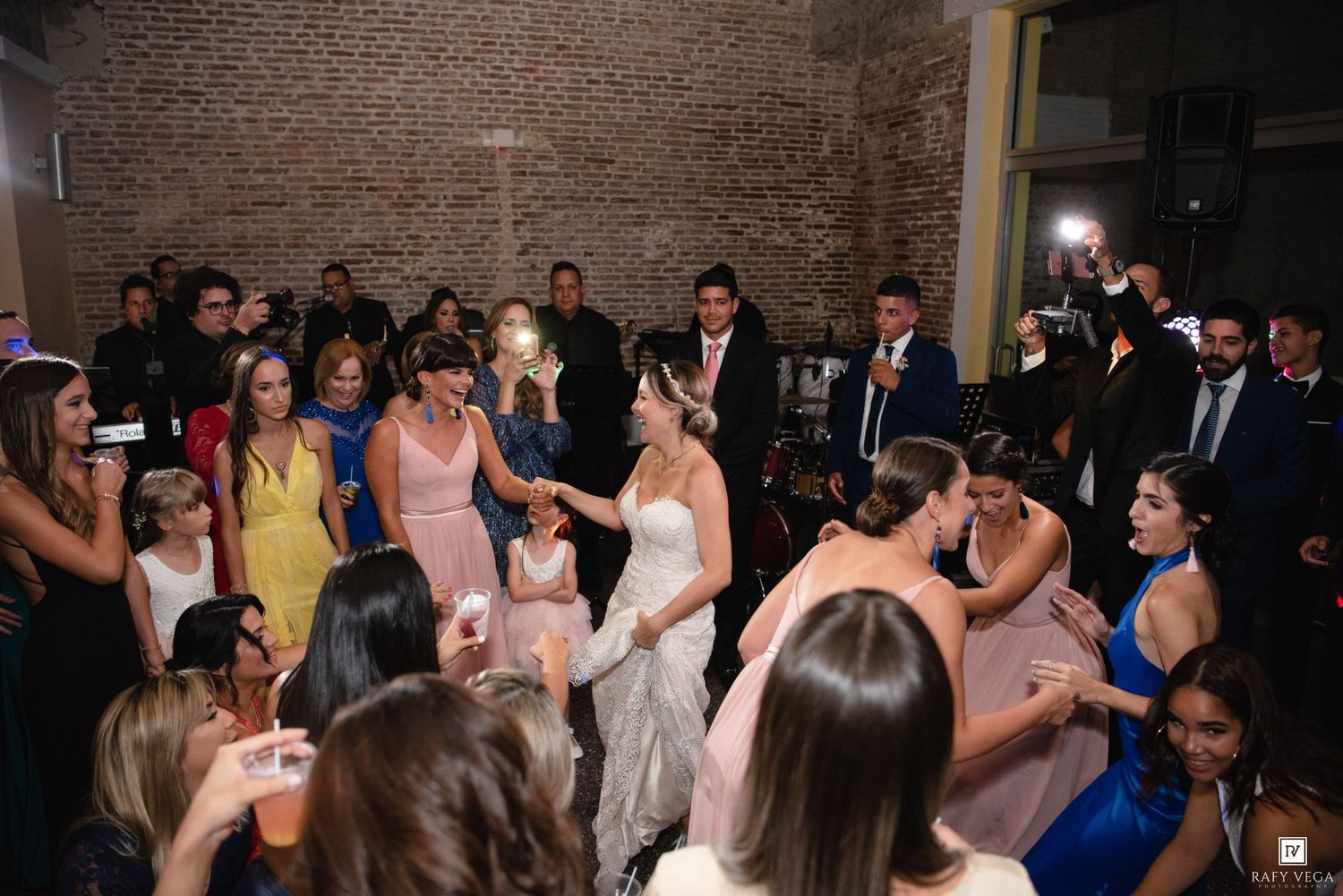 Bride dancing with guests at a lively wedding reception in a rustic brick-walled venue.