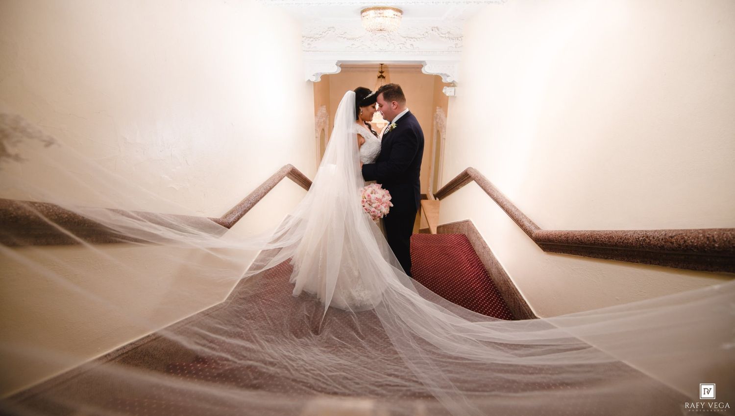 Bride and groom share romantic moment on grand staircase, cathedral veil flowing dramatically in elegant wedding venue.