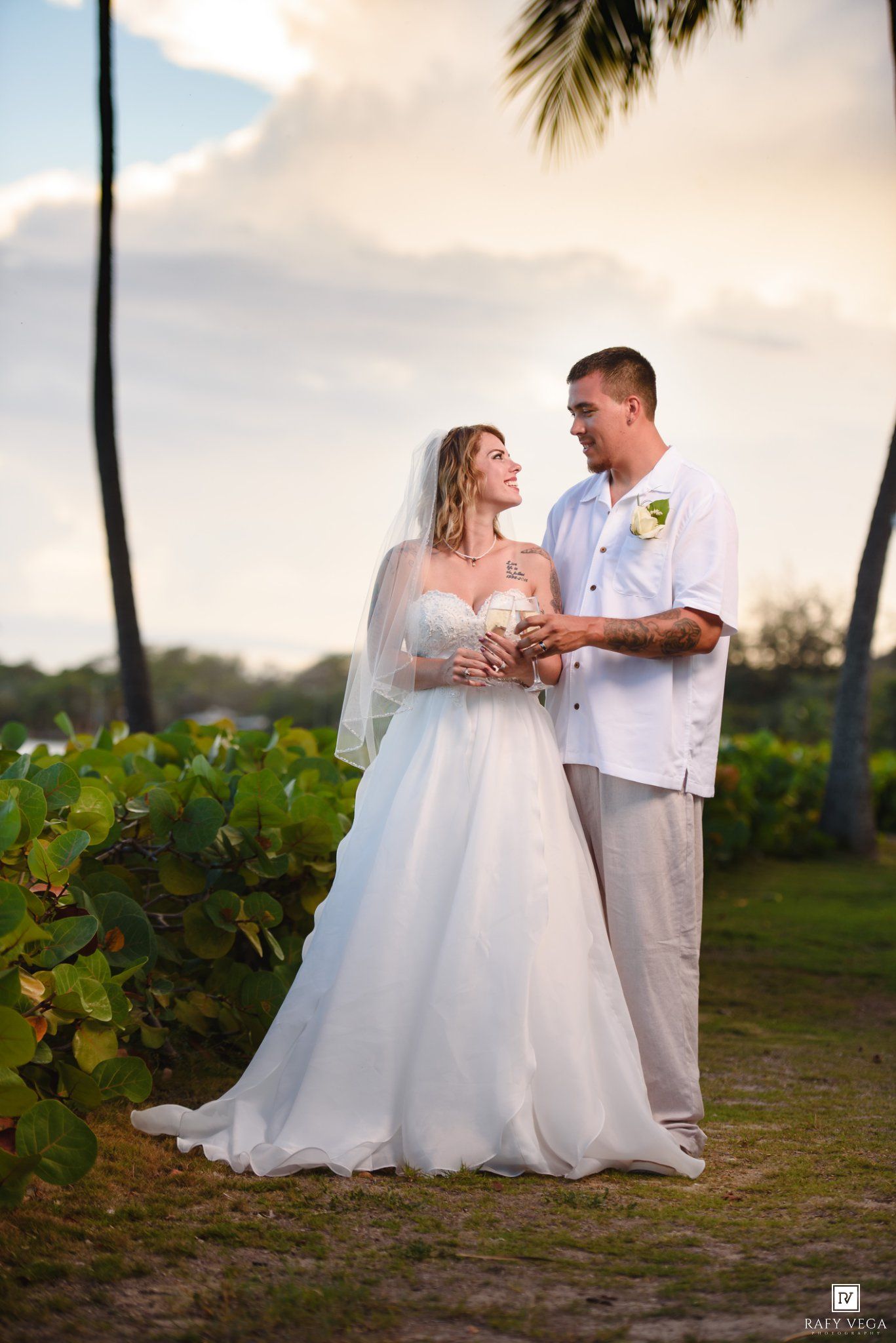 Bride in white gown and veil with groom in white shirt at tropical beach wedding ceremony.