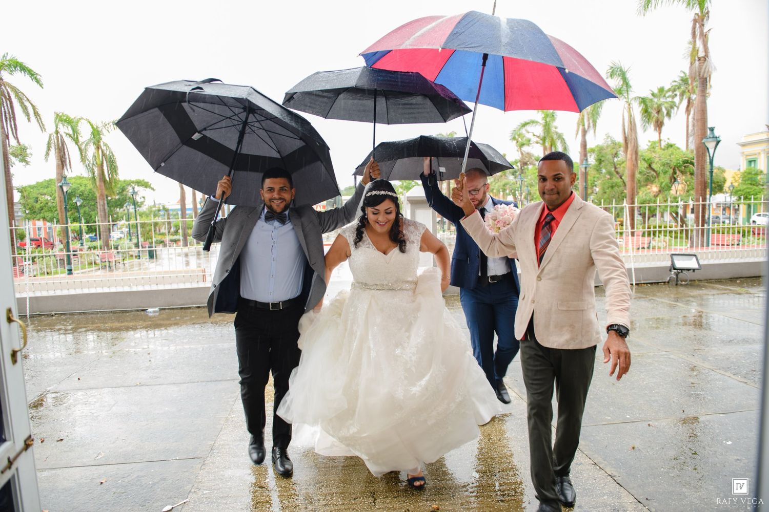 Bride in white gown with three groomsmen holding black and colorful umbrellas in the rain at outdoor wedding venue.