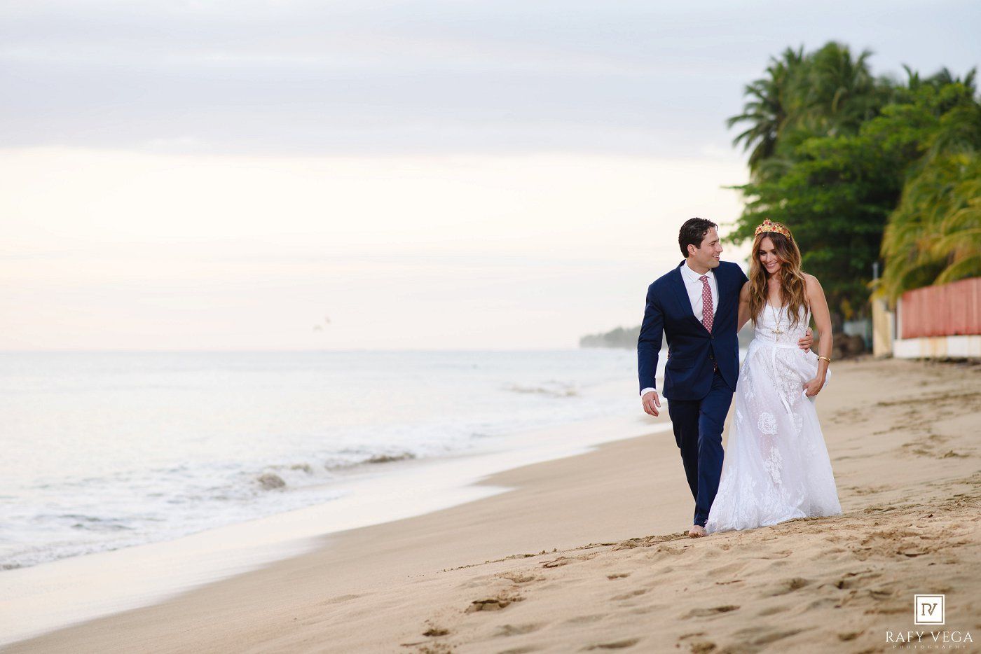 Bride and groom walking hand in hand on a tropical beach at sunset in wedding attire.