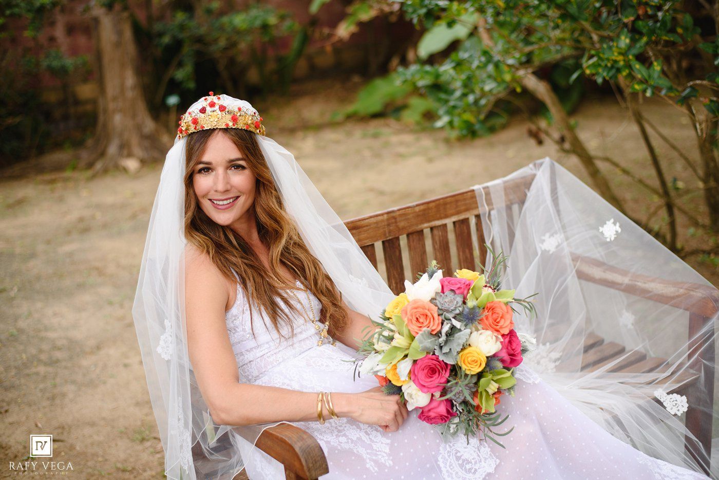 Smiling bride in white gown and floral crown holds colorful bouquet while seated on wooden bench outdoors.
