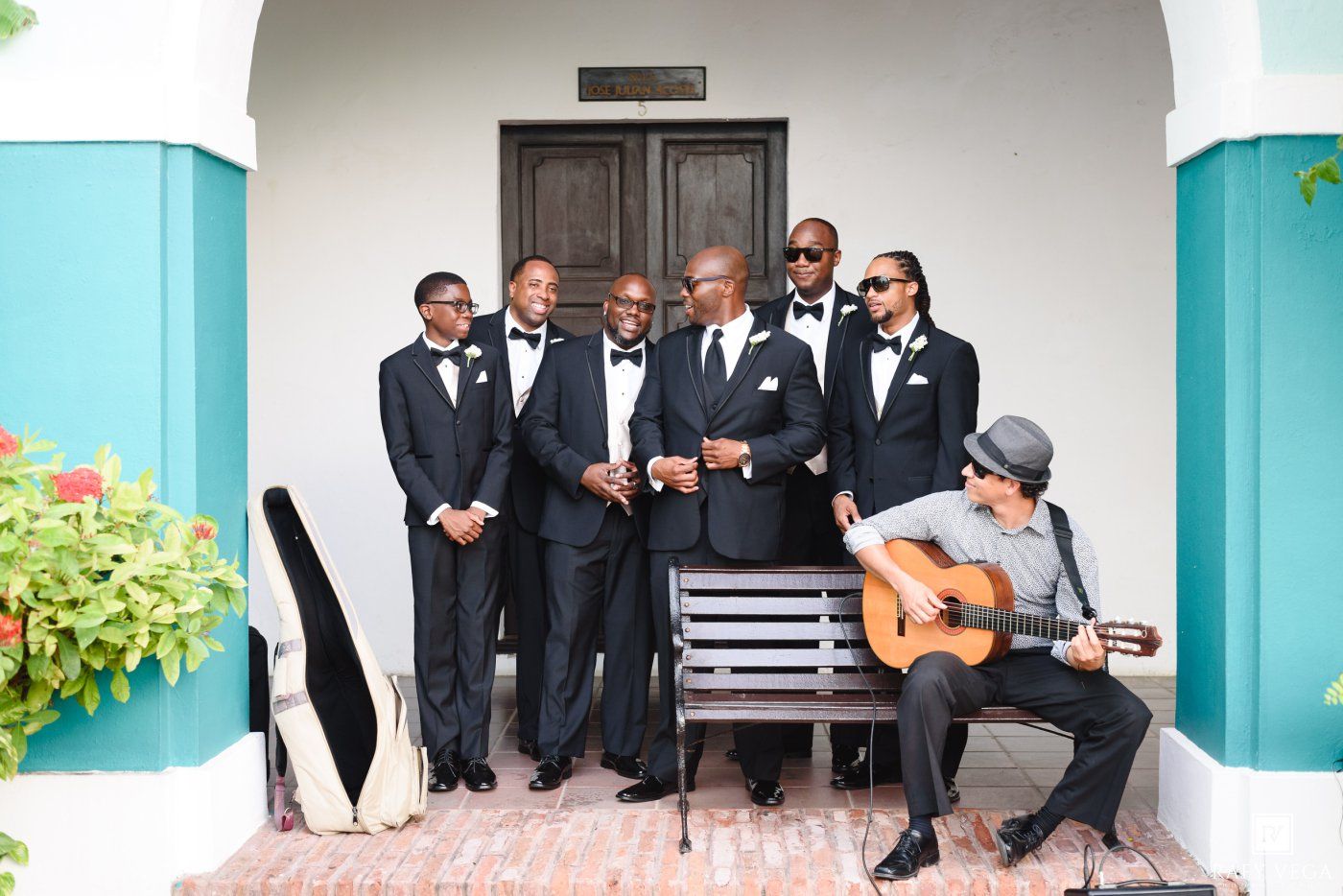 Groomsmen in tuxedos pose with guitarist at elegant venue entrance during outdoor wedding celebration.