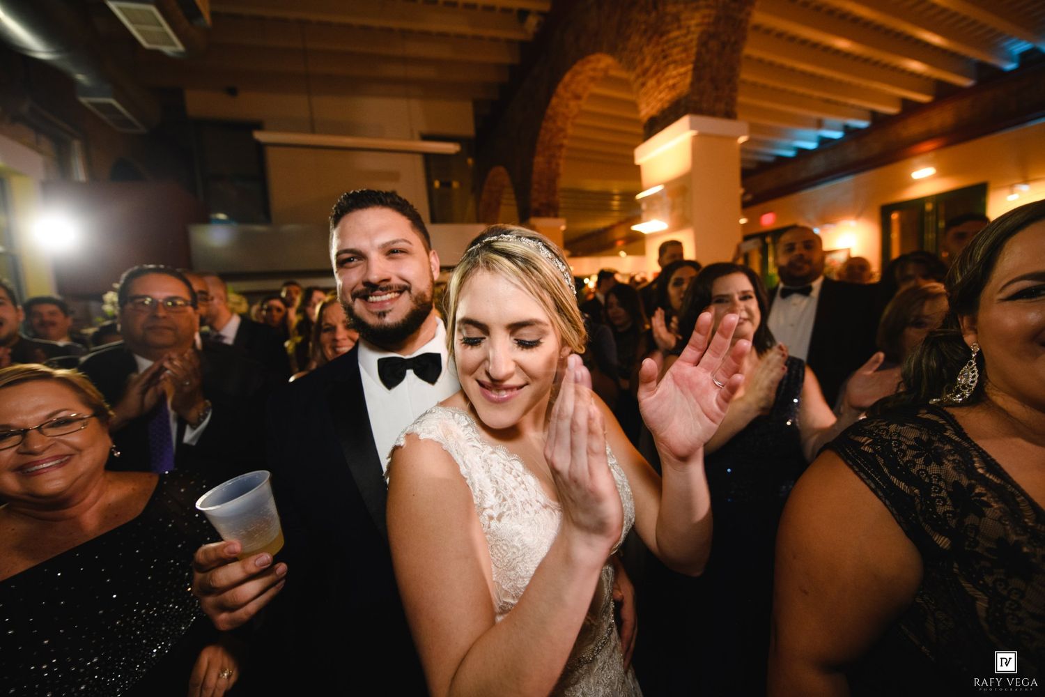 Bride and groom clapping and smiling joyfully at their wedding reception surrounded by guests in elegant attire.