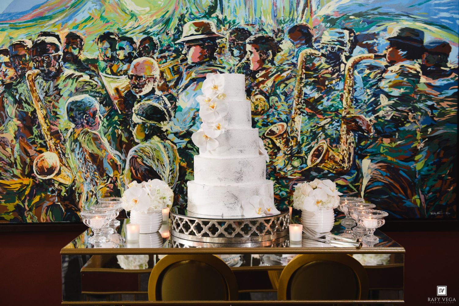 Elegant white wedding cake on gold table before vibrant jazz musician mural at upscale venue.