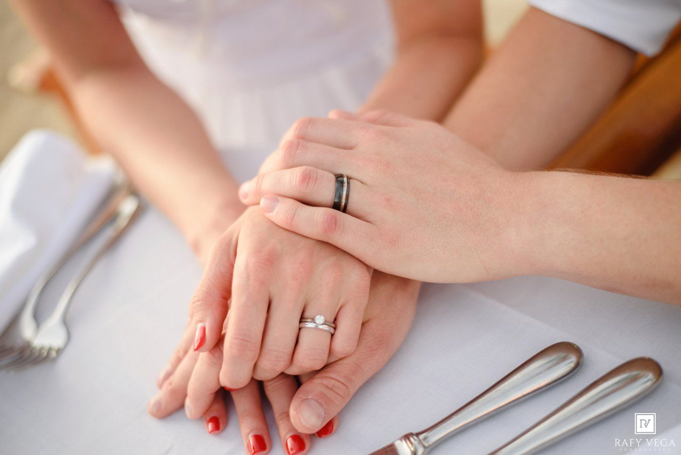 Newlywed couple displaying wedding rings on hands placed together on white table with silverware nearby.