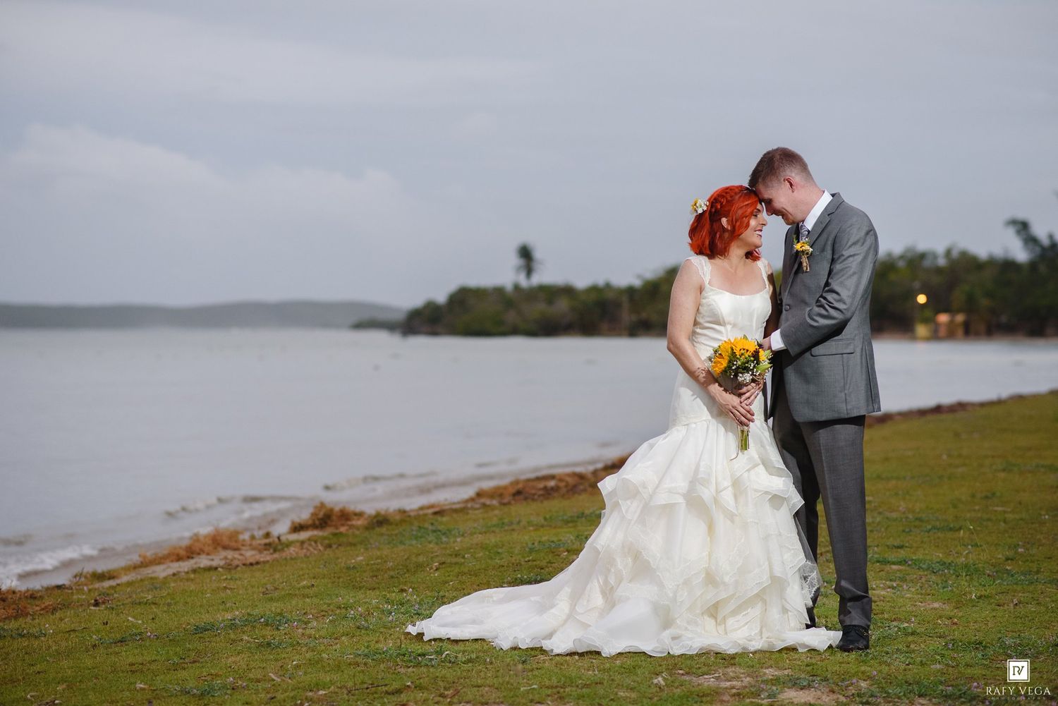 Bride in white gown and groom in gray suit share romantic moment by waterfront on cloudy wedding day.