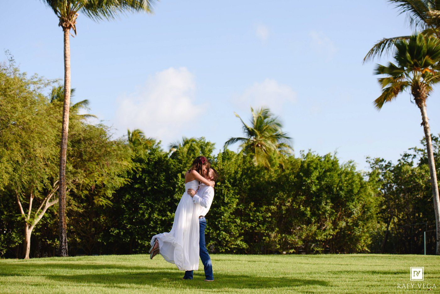 Couple embracing on lush green lawn surrounded by tall palm trees under a bright blue sky in a tropical setting.
