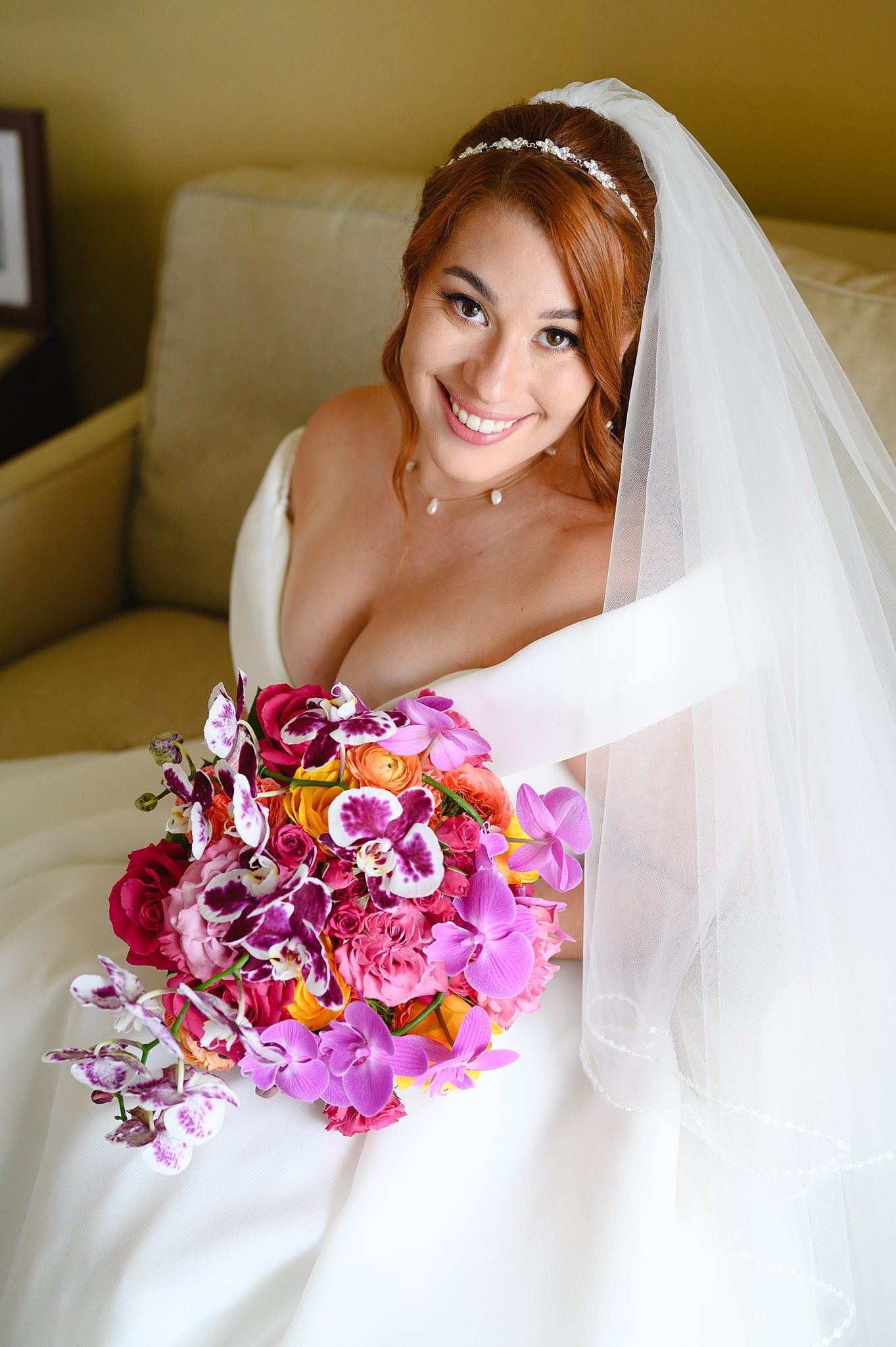 Smiling bride in white gown and veil holds vibrant pink, purple, and orange floral bouquet while seated on a couch.