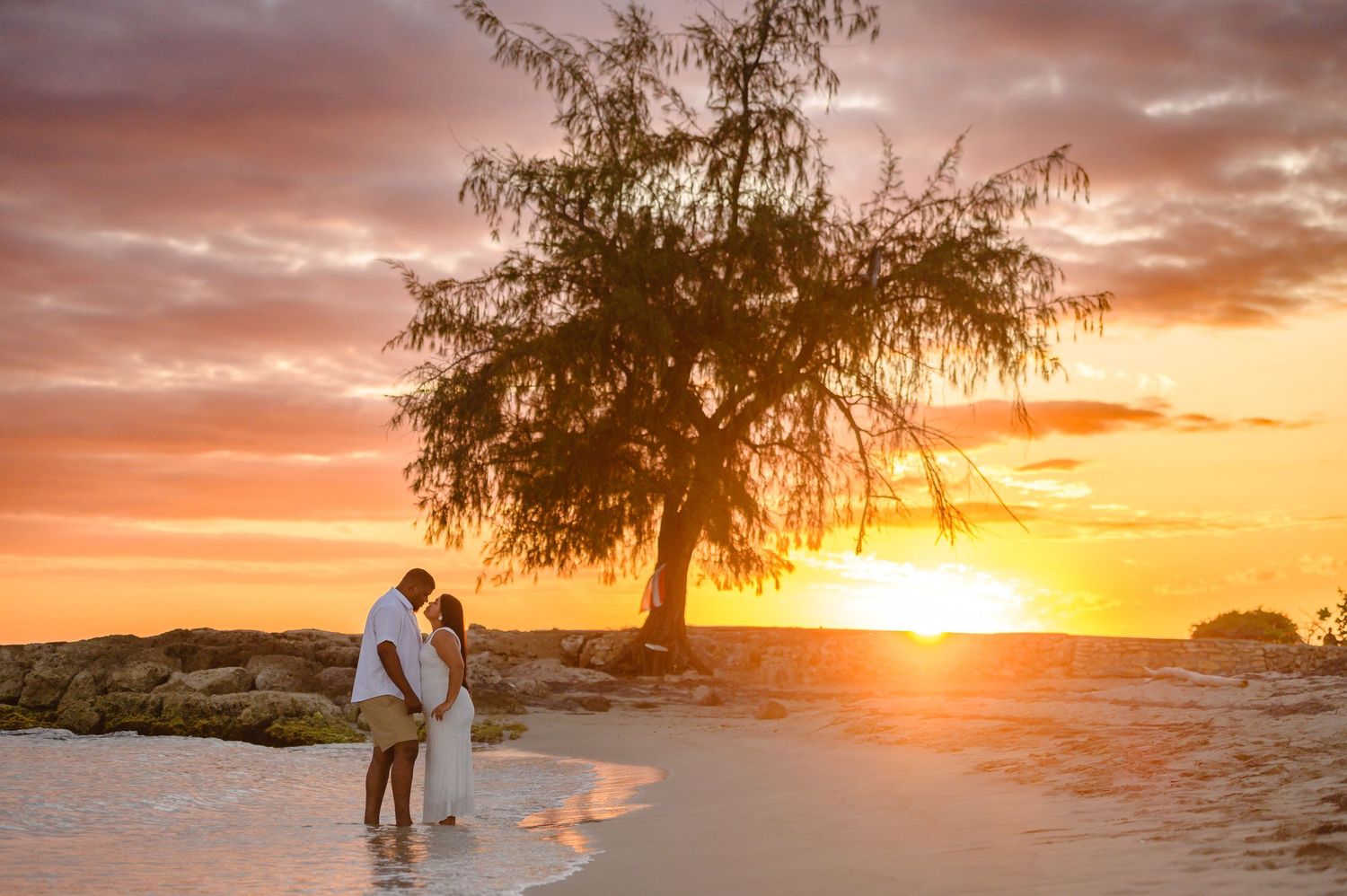 Couple embracing on a tropical beach at golden sunset with a silhouetted tree and dramatic orange sky.
