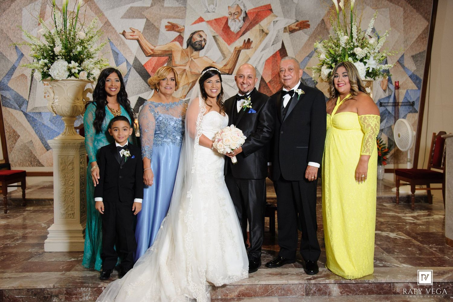 Wedding party poses in church with bride in white gown, groom in tuxedo, and guests in blue and yellow formal attire.