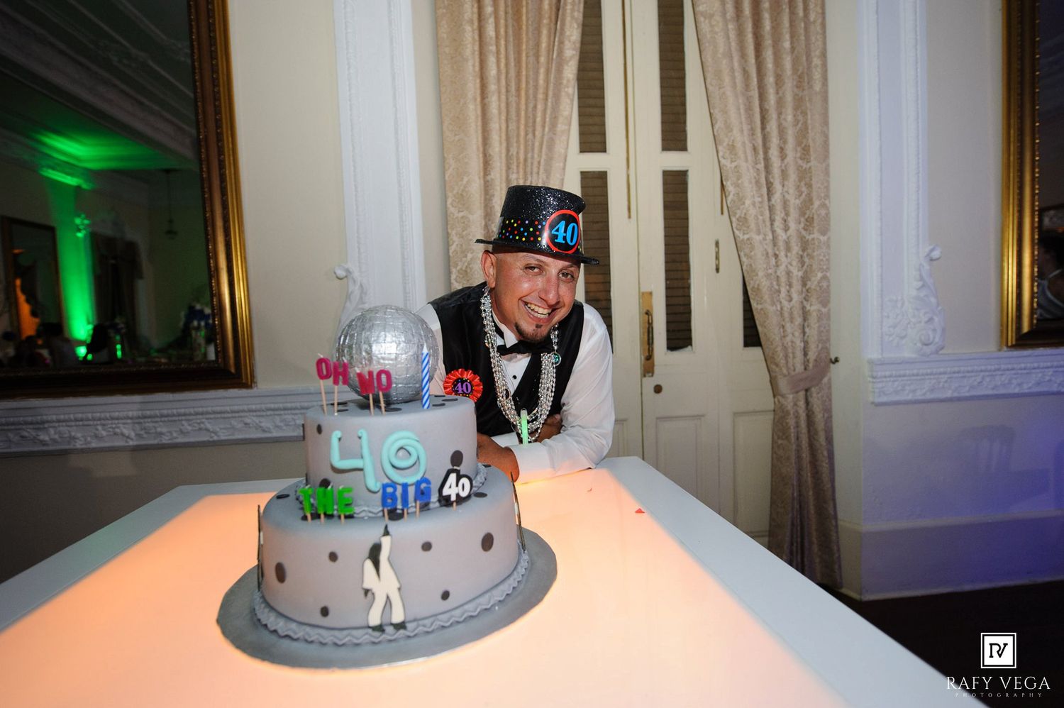 Man in top hat smiling behind a multi-tiered decorated birthday cake at an elegant indoor celebration.