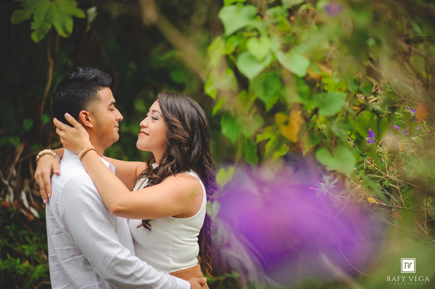 Couple embracing romantically outdoors surrounded by lush green foliage and purple flowers in a garden setting.