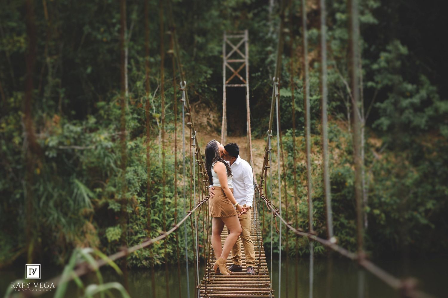 Couple sharing a romantic kiss on a wooden suspension bridge surrounded by lush green tropical forest.