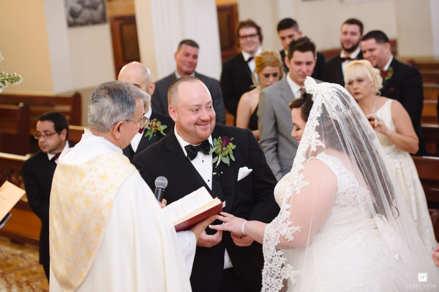 Bride and groom exchanging vows at church altar with officiant and wedding party in background.
