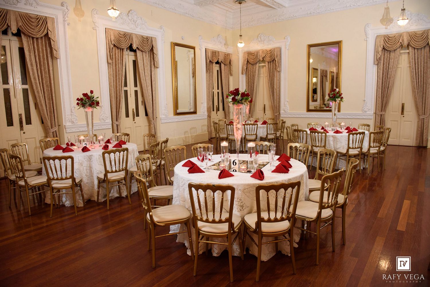 Elegant wedding reception hall with round tables, gold chairs, red floral centerpieces, and warm lighting on hardwood floors.