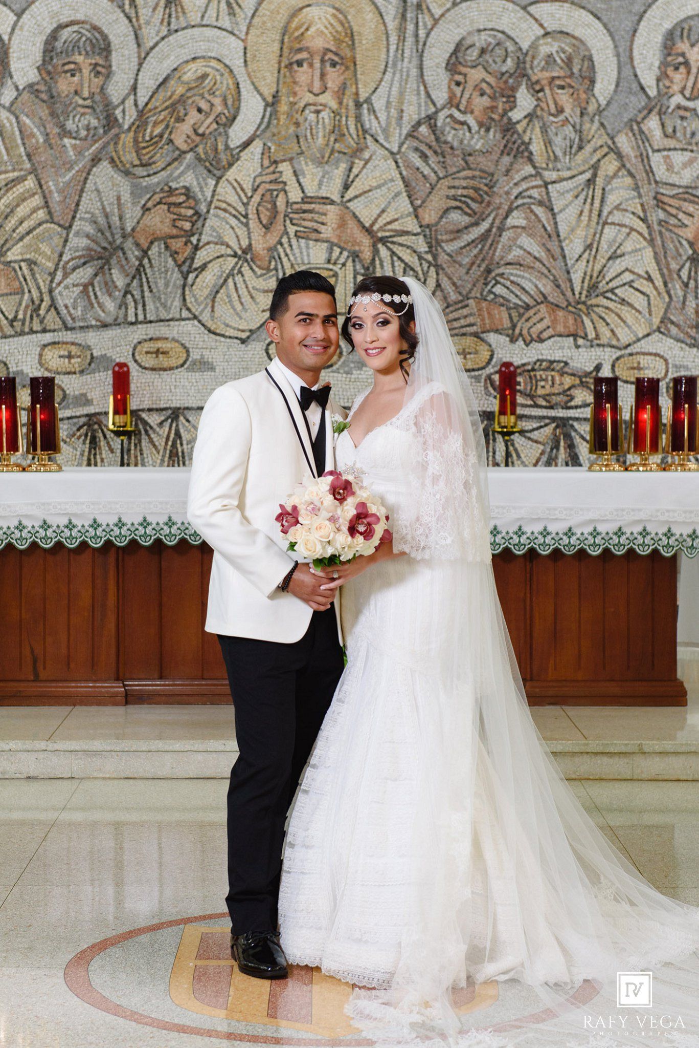 Bride and groom posing at church altar with mosaic mural backdrop, bride in lace gown holding bouquet.