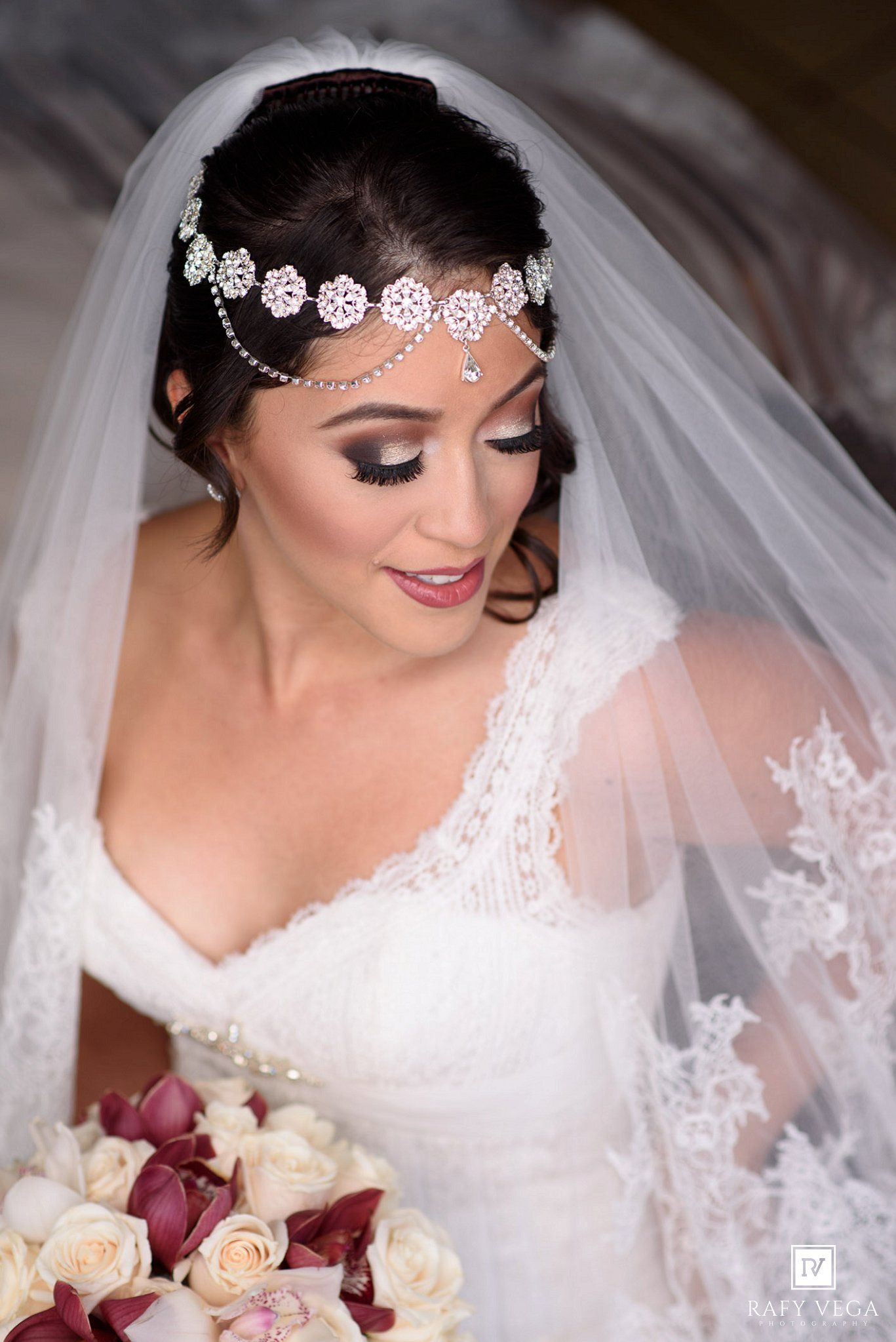 Elegant bride wearing a jeweled headpiece, veil, and lace gown, holding a bouquet of cream and burgundy roses.