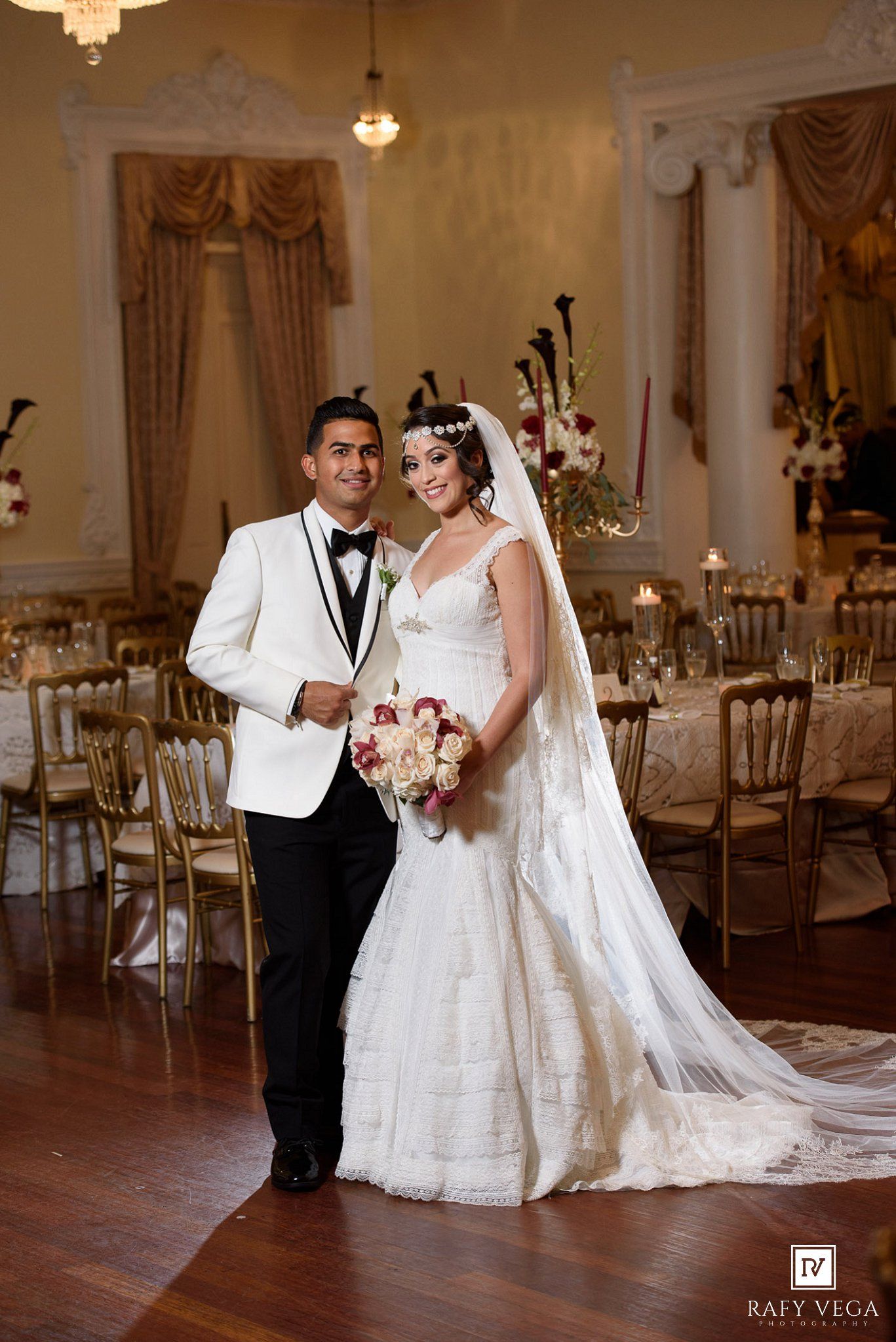 Bride and groom posing in ornate ballroom, groom in white tuxedo, bride in lace gown holding bouquet.