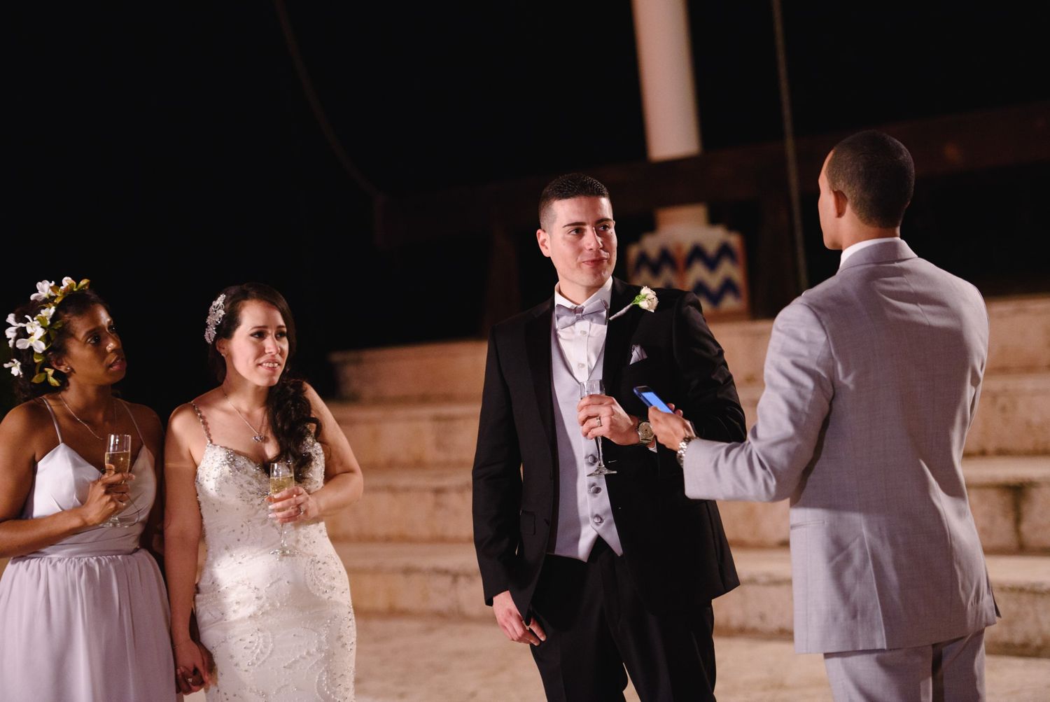 A groom in a black suit listens as a man in gray speaks at an outdoor wedding ceremony with bridesmaids nearby.