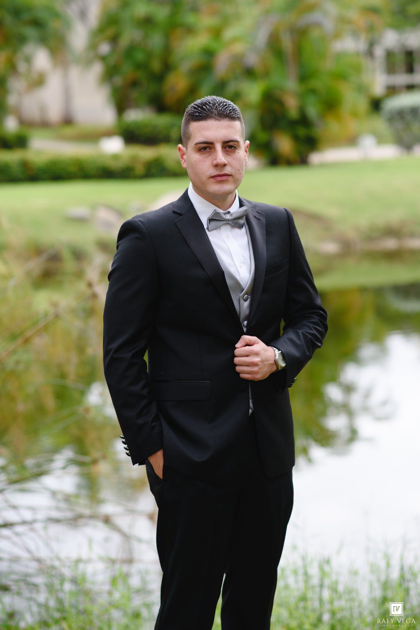 Elegant groom in black tuxedo with gray bow tie posing outdoors near a pond at wedding venue.