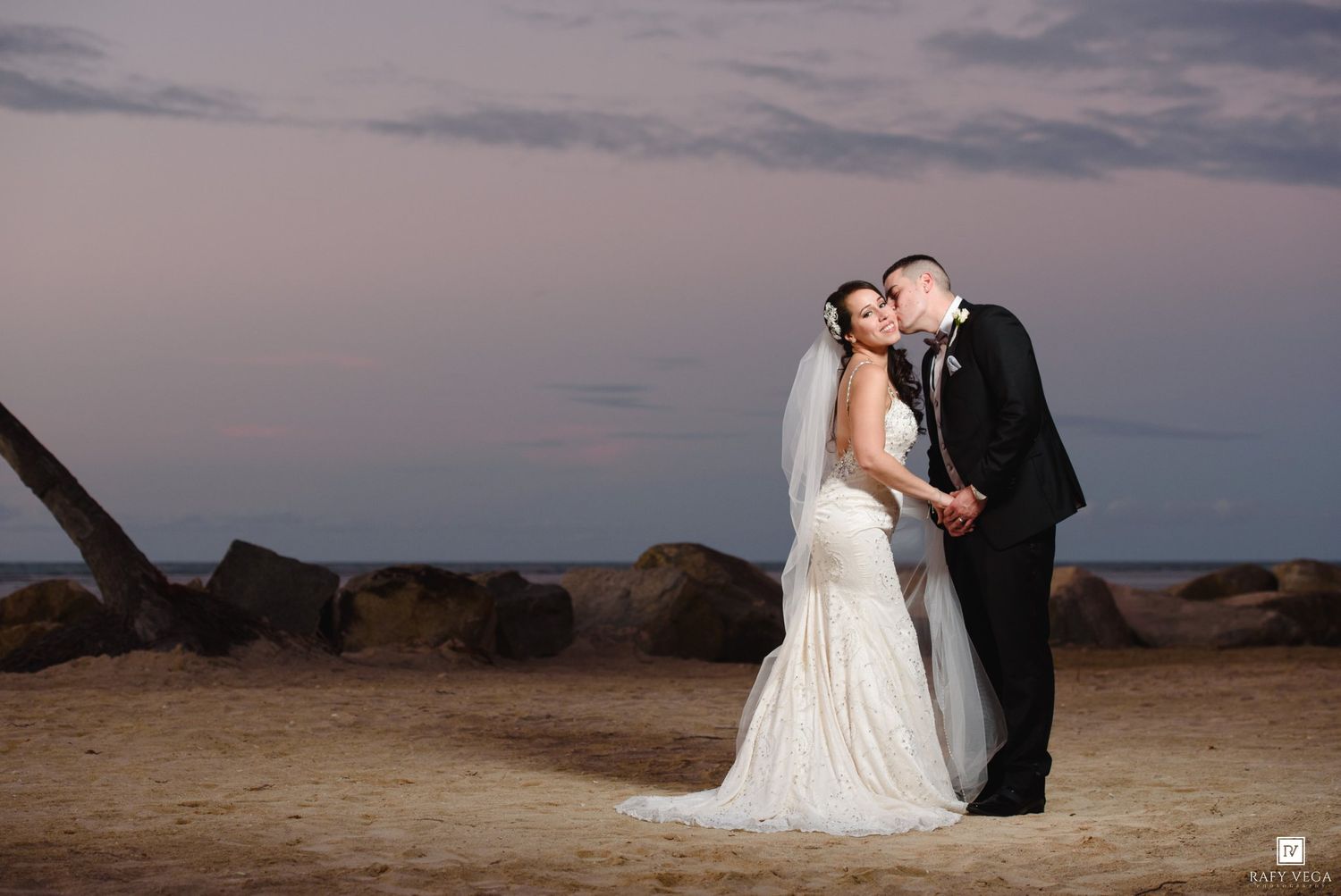 Bride and groom share a romantic kiss on a sandy beach at sunset, with rocks and pastel sky in the background.
