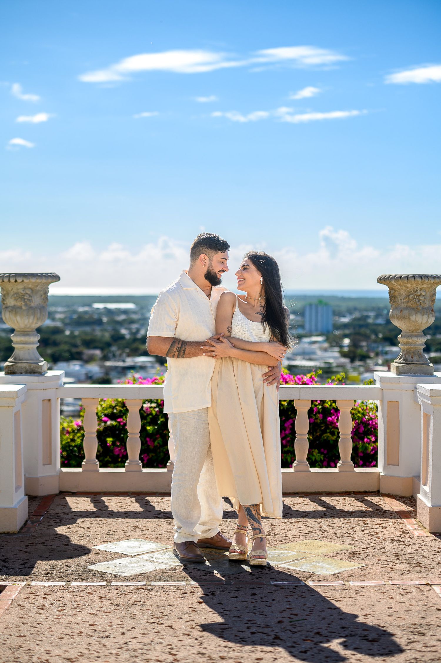 Couple embracing on elegant white balcony with colorful flowers and city skyline under blue sky.