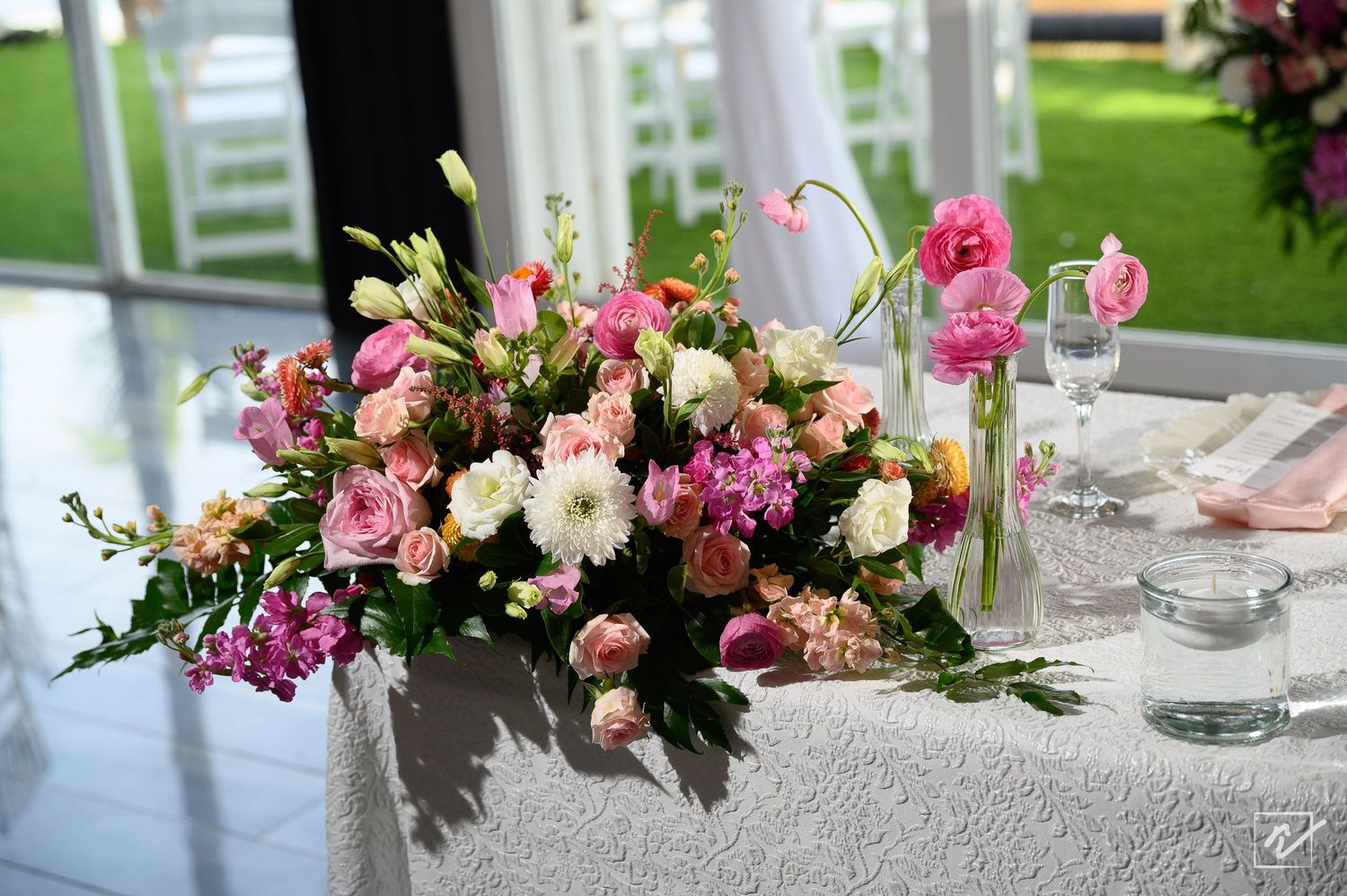 Pink, coral, and white floral centerpiece arrangement with roses and greenery on a white-draped wedding reception table with glassware.