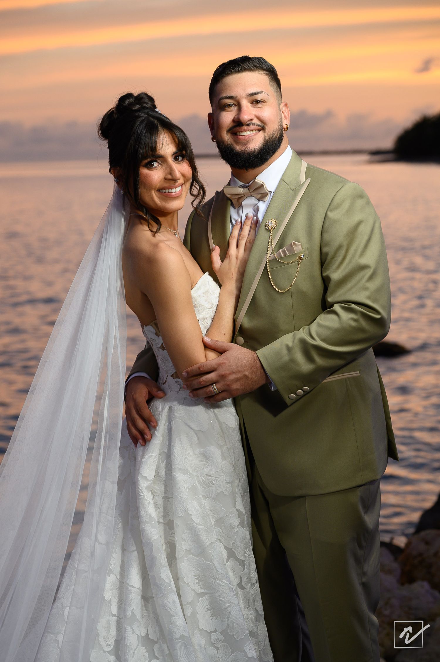Smiling bride in a lace gown and long veil embraces her groom in an olive green suit and bow tie on a waterfront at sunset.