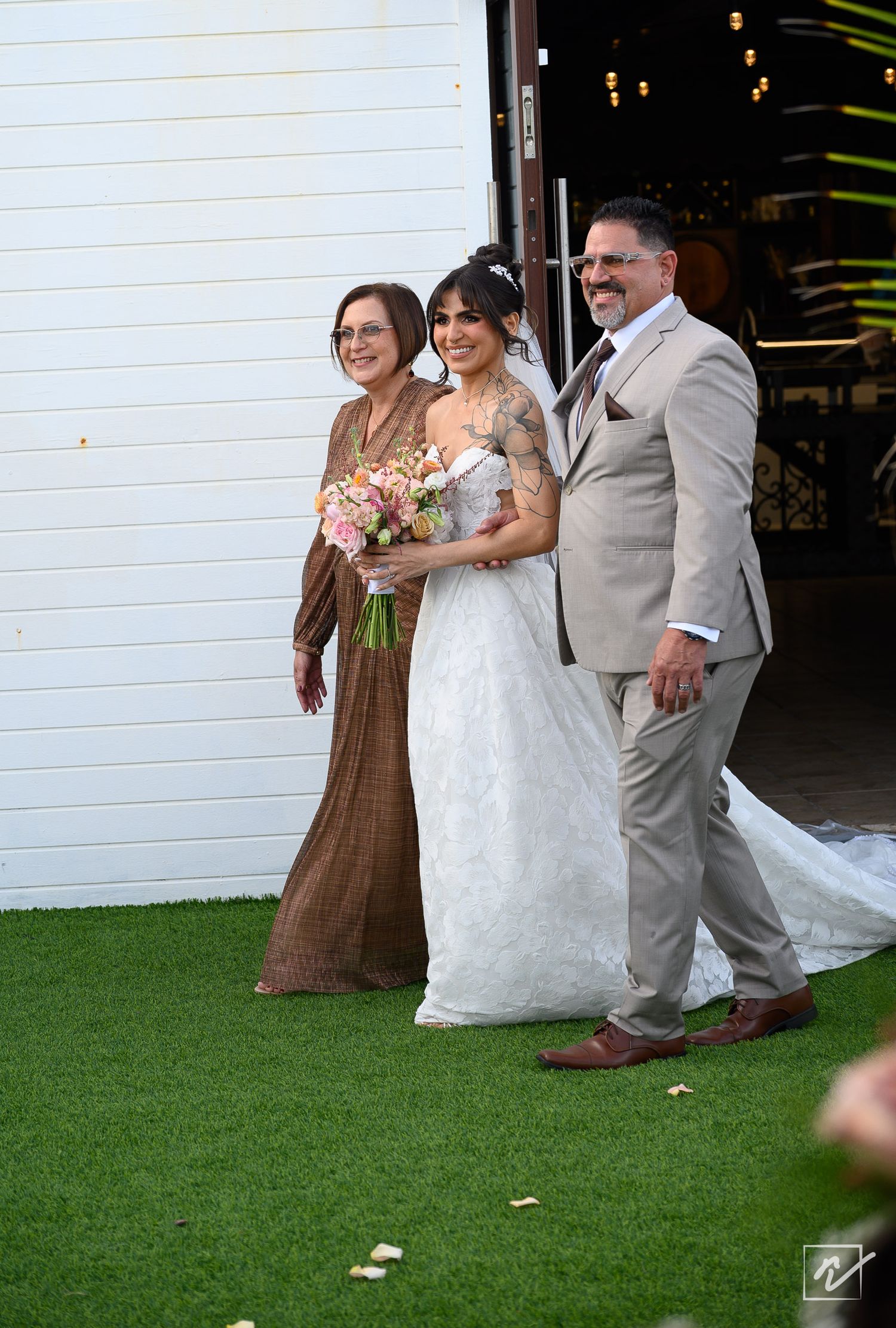 A bride in a white lace gown holding a pink bouquet walks on green grass with a man in a gray suit and a woman in a brown dress.