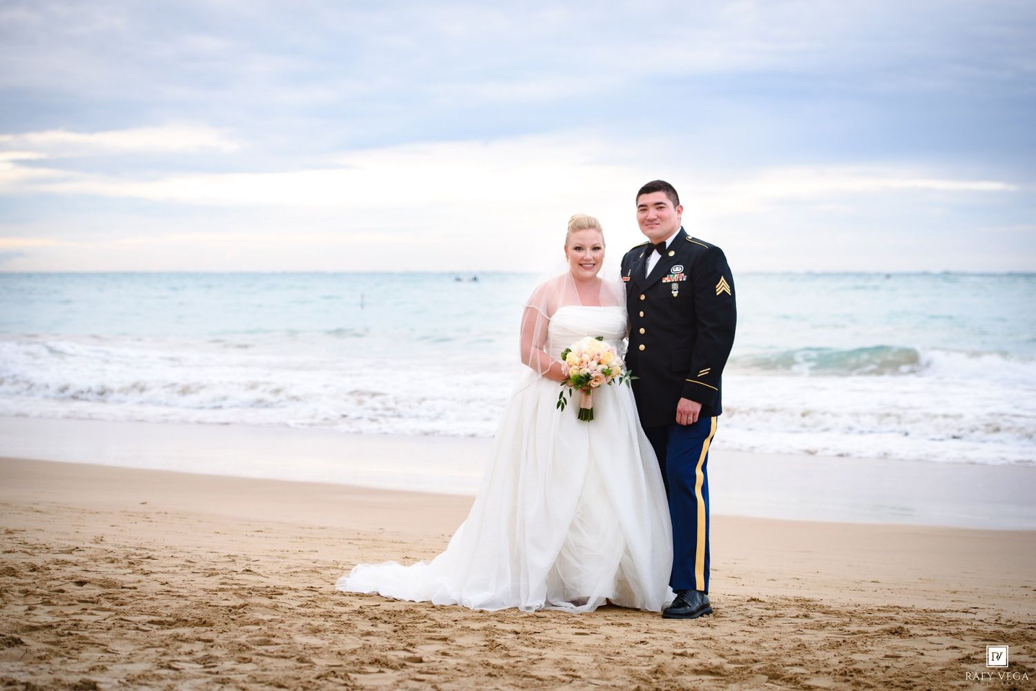 Military groom in dress uniform stands with bride in white gown holding bouquet on sandy beach at sunset.