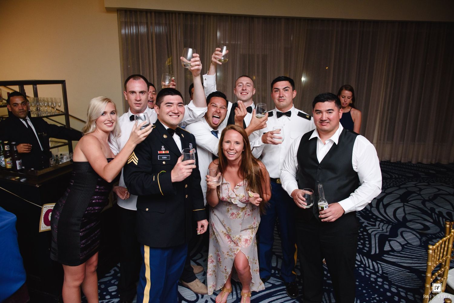 Group of wedding guests in formal attire raising glasses for a celebratory toast at a reception.