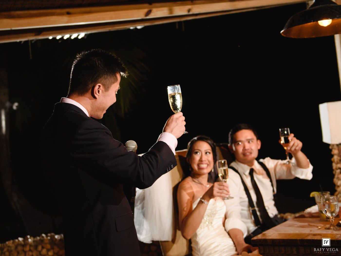 Bride and groom raising champagne glasses in a toast with guests at an elegant evening wedding reception.