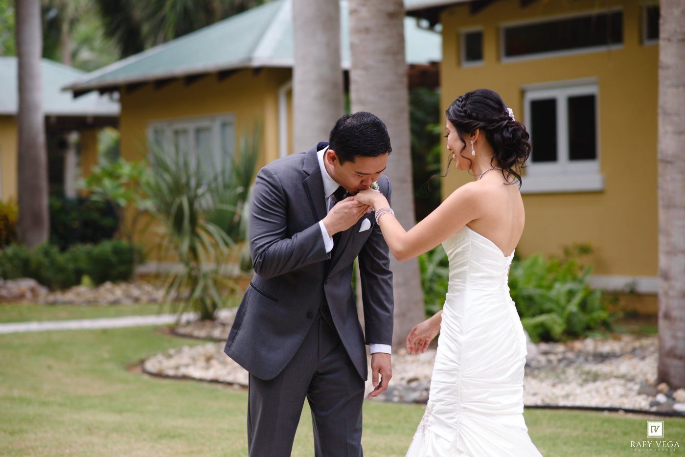 Groom kisses bride's hand during emotional outdoor wedding first look at tropical venue.