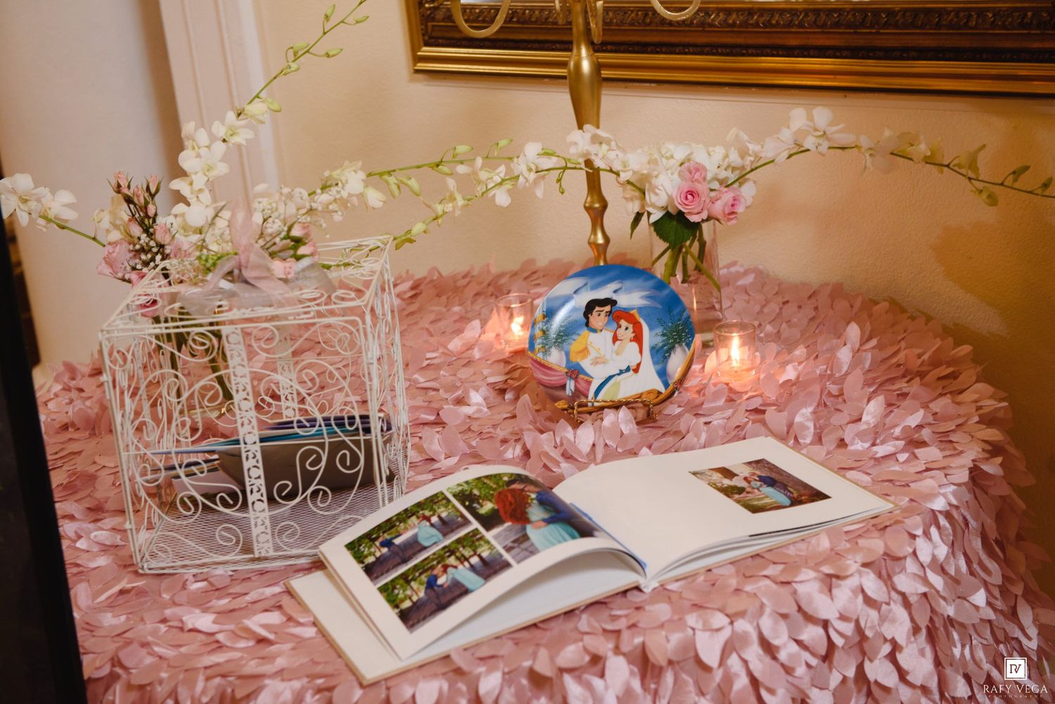 Pink ruffled table with open wedding photo album, white birdcage, floral vase, and decorative plate at a reception.