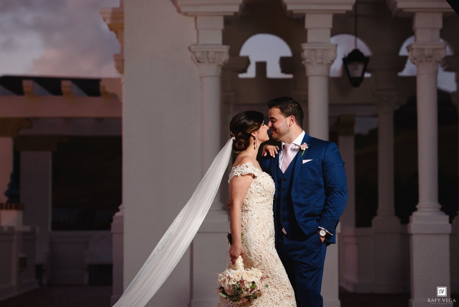Bride and groom share a romantic moment at dusk, with elegant white columns framing their wedding portrait.