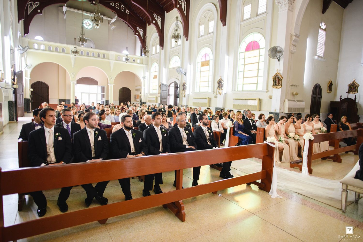 Elegant wedding ceremony in a grand church with groomsmen seated in wooden pews during a formal religious service.