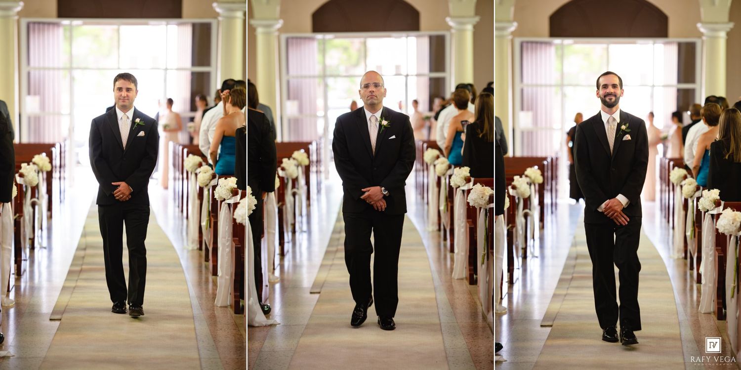 Groom stands at church altar as wedding party walks down aisle lined with floral arrangements during indoor ceremony.