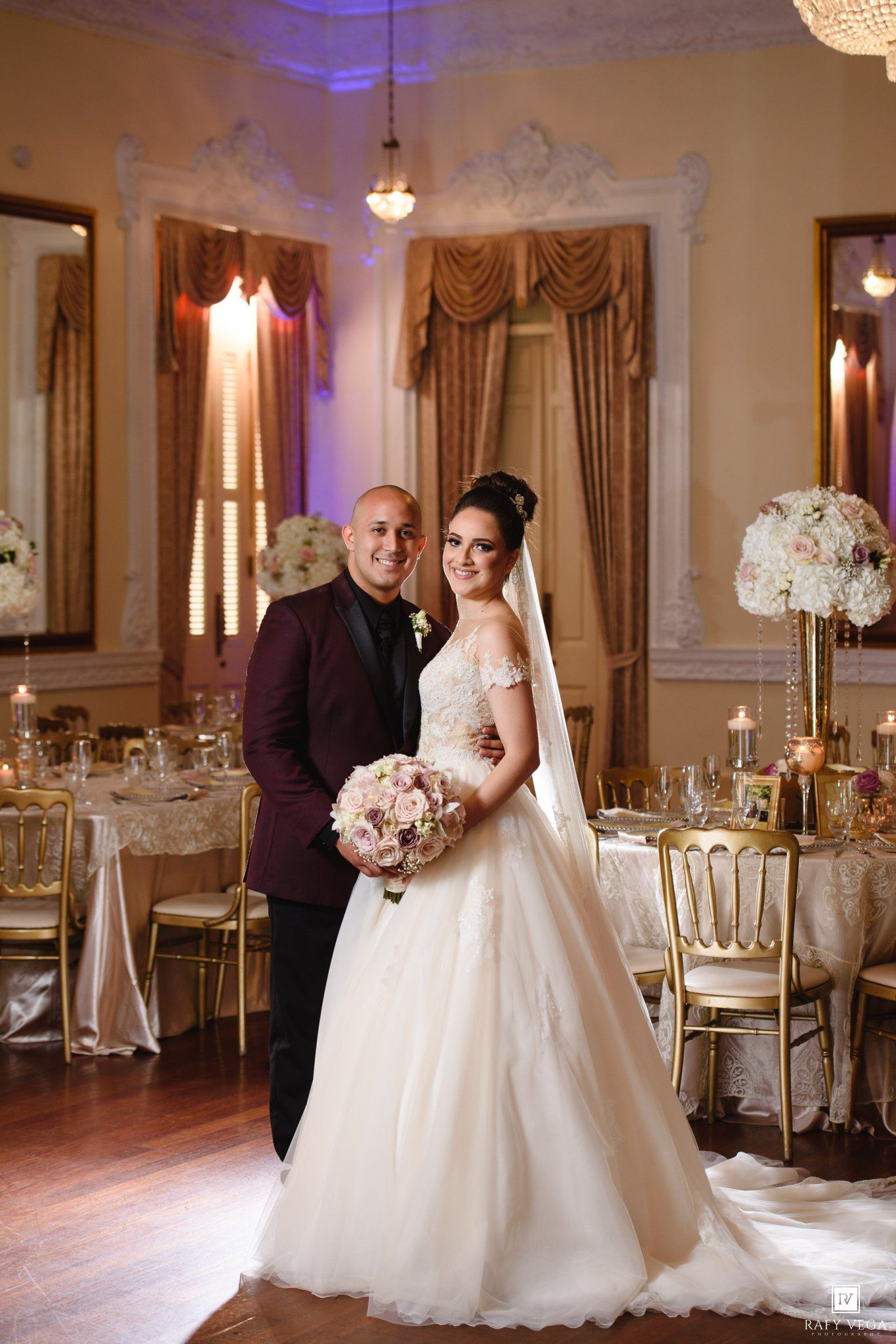 Bride and groom pose elegantly in a luxurious gold-decorated wedding reception hall, bride holding a pink bouquet.